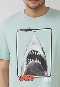 Tyrkysové bavlněné tričko s černobílou grafikou žraloka, siluetou plavce a výrazným červeným textem "JAWS 1975".