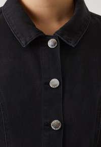 Veste en denim noir avec un col classique, des boutons-pression argentés et un tissu texturé. Gros plan sur les détails avant.