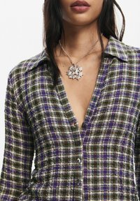 Chemise à carreaux avec boutons en vert, blanc et violet. Présente un col pointu et un tissu texturé. Associée à un collier argenté à motif d'étoiles.