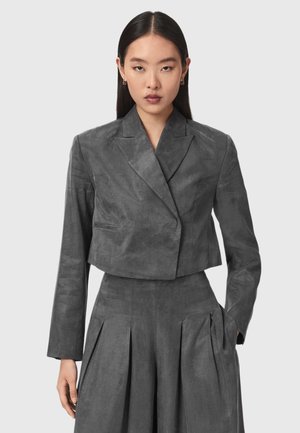 Vrouw draagt een op maat gemaakte, donkergrijze cropped blazer met lange mouwen en bijpassende hooggetailleerde geplooide broek op een neutrale achtergrond.