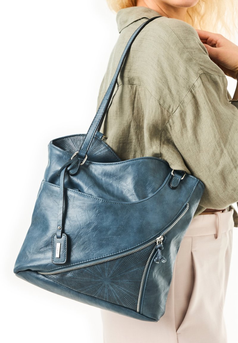 Sac fourre-tout en cuir bleu avec un design courbé, comportant un motif perforé et une fermeture éclair en argent. Comprend une bandoulière réglable et une étiquette.