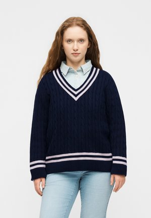 Femme aux longs cheveux roux portant un pull en maille torsadée bleu marine avec des rayures rose clair sur une chemise à col bleu clair et un jean bleu clair.