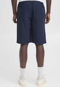 Tmavomodré ľanové šortky s elastickým pásom, dvoma zadnými vreckami a voľným strihom, ktoré končia tesne nad kolenami. Biele ponožky a topánky.