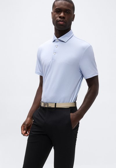 Άνδρας που φοράει ανοιχτό μπλε πόλο πουκάμισο με υφή, φορεμένο μέσα σε μαύρο παντελ όνι με μπεζ ζώνη, στέκεται με το ένα χέρι στην τσέπη μπροστά σε λευκό φόντο.