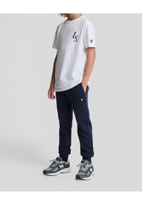 Witte katoenen T-shirt met "LS" logo op de linkerborst, gecombineerd met marineblauwe fleece joggingsbroek met een gele embleem. Draagt grijze sneakers.