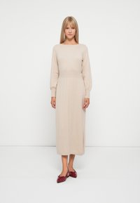 Abito beige in maglia con maniche lunghe, scollatura a barca e vita aderente. Presenta un design di lunghezza media e una texture sottile. Abbinato a scarpe rosse.