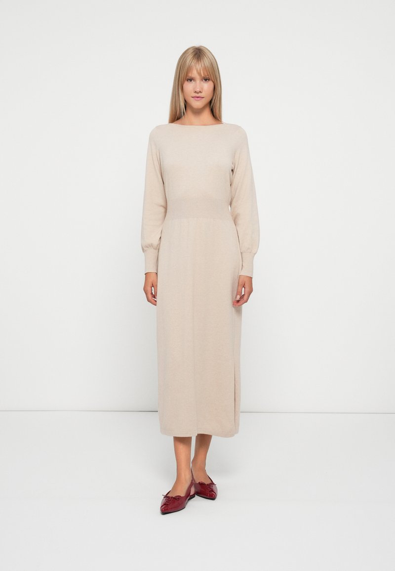 Abito beige in maglia con maniche lunghe, scollatura a barca e vita aderente. Presenta un design di lunghezza media e una texture sottile. Abbinato a scarpe rosse.