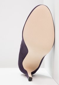 Chaussure à talon haut en suede violet avec un bout pointu et un talon aiguille fin. La semelle est beige avec des détails de grip texturé.