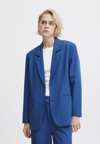 Blazer azul hecho de una tela suave, con solapas muestradas, dos botones y bolsillos laterales. El diseño es ajustado con un corte relajado.