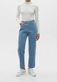 Jean en velours côtelé bleu clair taille haute avec une coupe droite, associé à un col roulé crème côtelé et des baskets blanches.