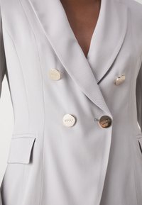 Blazer doppiopetto in tessuto grigio chiaro con revers a punta, due tasche e bottoni con logo in tono dorato. Tessuto di texture liscia.