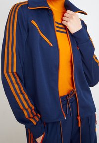 Marinblå träningsdräkt med orange detaljer, en jacka med dragkedja och hög krage samt randig design på ärmarna. Mjuk, texturerad material.