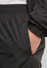 Veste coupe-vent noire en tissu léger texturé. Les poignets et la taille élastiques assurent un ajustement parfait. Poche latérale visible.