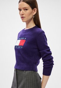 Μωβ πλεχτό πουλόβερ με ριχτές μανσέτες και στρίφωμα. Διαθέτει κολλητική ετικέτα με κόκκινες και λευκές λεπτομέρειες. Φοριέται με γκρι παντελόνι με ρίγες.