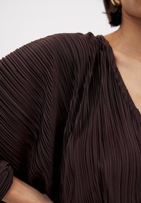 Tessuto di un abito marrone plissettato con scollatura a V profonda e vestibilità morbida, che mostra sottili pieghe verticali e una texture liscia.