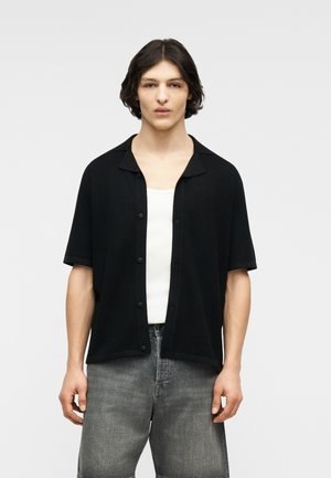 Jeune personne aux cheveux mi-longs et foncés portant une chemise noire à manches courtes boutonnée sur un débardeur blanc et un jean gris délavé.