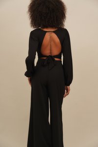 Black backless top with long sleeves and bow tie at the waist, paired with wide-leg black pants. Textured fabric with a smooth finish.