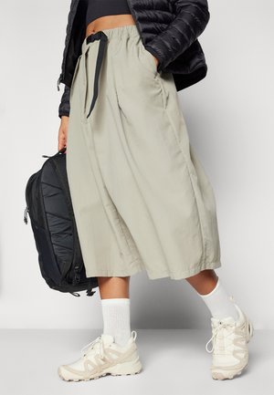 Personne portant un pantalon beige à jambes larges, des chaussettes blanches, des baskets blanches, une veste noire courte, et tenant un sac à dos noir à côté d'elle.