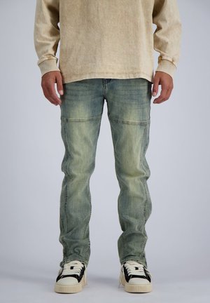 Mann trägt ausgewaschene grüne Jeans und beige Langarmshirt, steht mit leicht gespreizten Beinen und weißen Sneakers mit schwarzen Details.