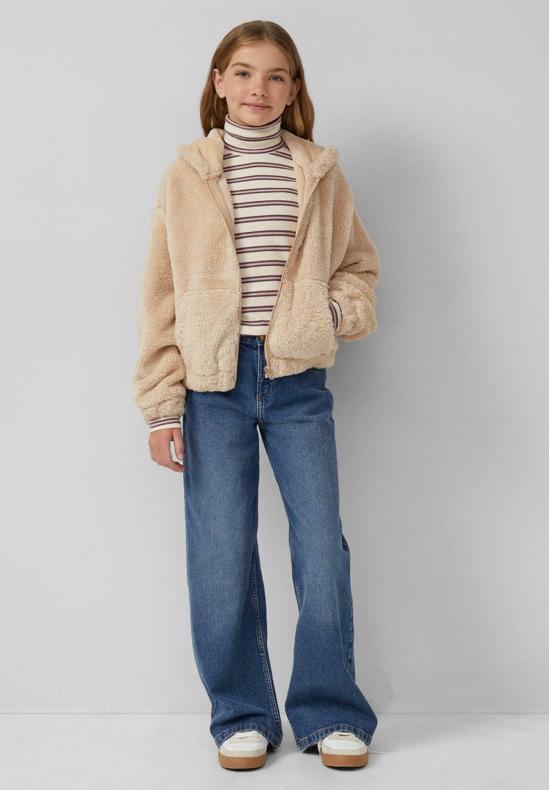 Junges Mädchen in einer beigen, flauschigen Zip-Jacke, gestreiftem Rollkragenpullover, weit geschnittenen blauen Jeans und weißen Sneakers, das vor einer grauen Wand steht.