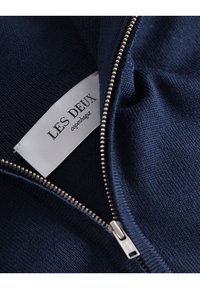 Tengerészkék kötött ruhadarab ezüst színű fémléccsel, részben nyitott állapotban, amely mögött egy fehér szövetcímke látható, rajta a "LES DEUX copenhague" felirattal.