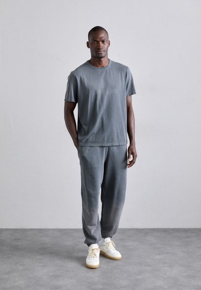Tricou gri din bumbac cu mânecă scurtă și guler rotund, asociat cu pantaloni de trening asortați. Adidași albi cu tălpi bej. Design simplu și minimalist.