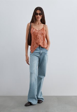 Mujer con un top sin mangas naranja con lentejuelas, vaqueros de pierna ancha azul claro, gafas de sol oscuras y un bolso de hombro, de pie frente a un fondo liso.