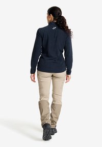 Frau mit dunklen Haaren im Pferdeschwanz, die eine marineblaue Jacke und beige Cargo-Hosen trägt, geht vor einem weißen Hintergrund weg.