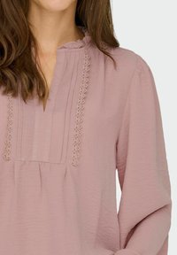Blusa rosa chiaro con scollatura a V, dettagli arricciati e righe verticali; tessuto testurizzato con una vestibilità morbida e dettagli in pizzo.