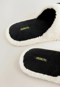 Černobílé plyšové pantofle s chlupatým bílým lemem, mají černou vnitřní výstelku a na každém pantofli zelenou etiketu "PEANUTS".