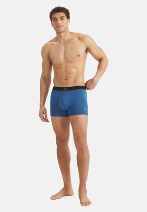 Man zonder shirt die de tailleband van blauwe boxershorts met zwarte tailleband en logo verstelt, staand op blote voeten tegen een witte achtergrond.