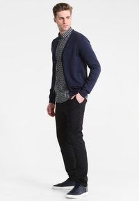 Marinblå cardigan över en rutigt skjorta, svarta byxor och marinblå sneakers. Cardigan har knappar, skjortan har ett litet rutmönster.
