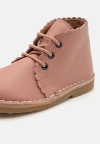 Botín de piel rosa con cuello ondulado, cordones marrones y ojales negros. Cuenta con costuras blancas y una suela marrón.