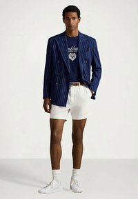 Άνδρας με ναυτικό ριγέ σακάκι, ναυτική μπλούζα με γραφικά, λευκά σορτς, λευκές κάλτσες και λευκά αθλητικά παπούτσια, στέκεται με το ένα χέρι στην τσέπη.