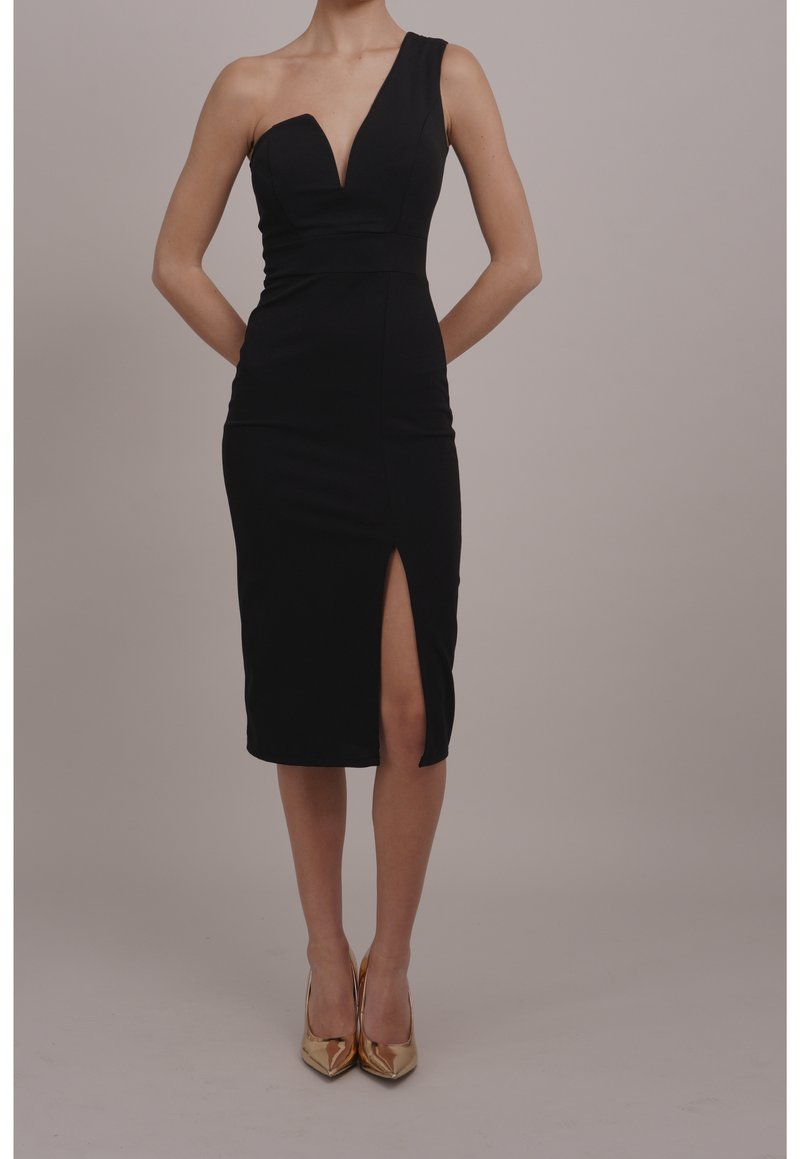 Femme portant une robe noire asymétrique longueur genou avec une fente à l'avant et des escarpins dorés à bout pointu, debout devant un fond uni.