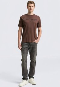 Camiseta unisex marrón con la inscripción "TOM TAILOR" en negro, combinada con vaqueros gris oscuro y zapatillas blancas, posando sobre un fondo neutro.