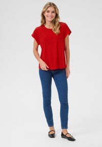 Haut rouge à manches courtes avec un design en œillet, associé à un jean skinny bleu et des ballerines noires, se tenant devant un fond clair.