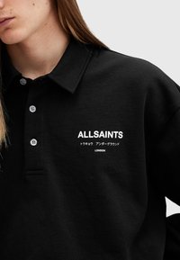 Musta polo-paita, jossa on kaulus ja valkoinen bränditeksti "ALLSAINTS" sekä lisäteksti japaniksi. Pehmeä, teksturoitu kangas, jossa on napit.