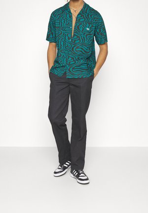 Mann trägt ein kurzärmeliges Hemd mit blauem und schwarzem Zebramuster, schwarze Hose und schwarz-weiße Turnschuhe, steht mit den Händen in den Taschen.