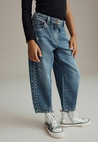 Enfant portant un jean bleu ample avec des détails cloutés et des baskets montantes argentées avec lacets et fermetures éclair latérales, debout devant un fond uni.