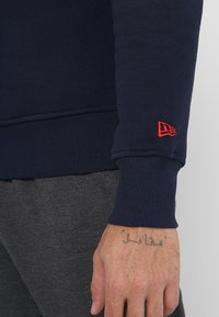 Sudadera de color azul marino con puños y dobladillo acanalados. Presenta un pequeño logo bordado en rojo en la manga izquierda. Tejido suave y texturizado.