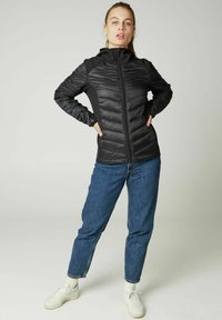 Zwarte gewatteerde jas met een voorkant rits en capuchon, voorzien van een gladde textuur en contrasterende zijpanelen. Gecombineerd met blauwe jeans en witte sneakers.