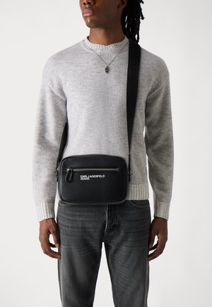 Borsa a tracolla nera con tasca con zip, finitura testurizzata e logo. Indossata su un maglione grigio e abbinata a jeans scuri. Sfondo semplice.