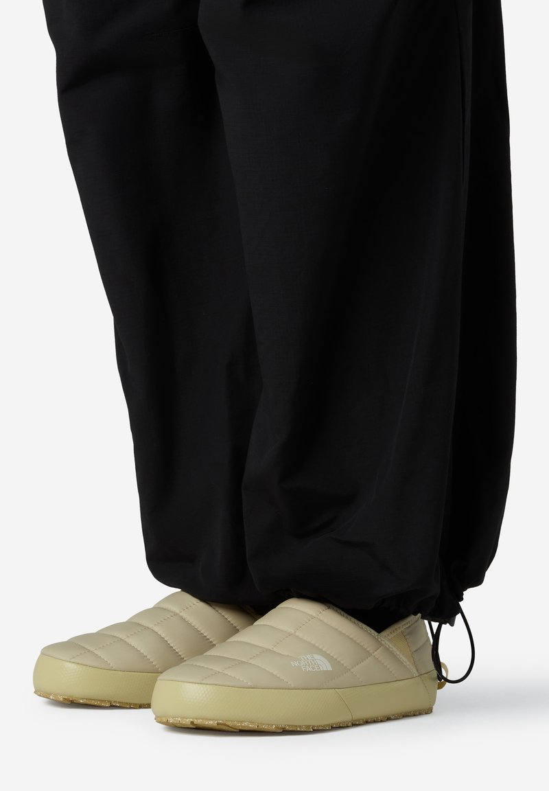 Zapatillas de slip-on acolchadas en color beige con una superficie texturizada y un logo, combinadas con pantalones negros de corte suelto que presentan elástico en los tobillos.