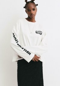 Bílá košile s dlouhým rukávem s černou grafikou "Stranger Things" na hrudi a nápisem "HELLO THERE LIVES" tištěným vertikálně na rukávu.