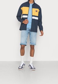 Muž oblečený v námořnické, žluté a bílé bundě Tommy Jeans přes modrou košili, světlých džínových šortkách, bílých ponožkách a bílých teniskách s námořnickými akcenty.