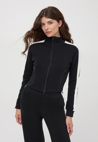 Czarna kurtka zapinana na zamek z wysokim kołnierzem i dwiema kieszeniami na zamek. Białe paski na ramionach. Gładka tekstura, dopasowany krój, krótka długość.