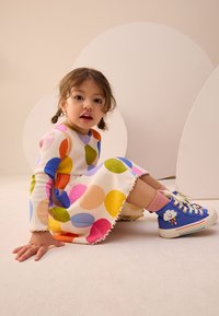 Robe colorée à pois avec manches longues, ourlet festonné et teintes vibrantes. Baskets bleues avec un motif de nuage et des rayures, chaussettes roses aux chevilles.