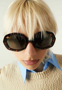 Lunettes de soleil octogonales avec une monture en écaille noire et des verres noirs, présentant des branches épaisses et une finition lisse. Un pull en tricot de couleur claire est visible.