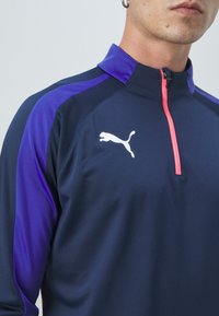 Jūrų laivyno ir violetinės spalvos sportinė ketvirčio užtrauktuko palaidinė, su rožine užtrauktuku ir baltu Puma logotipu priekyje. Pagaminta iš lengvos, tekstūruotos medžiagos.